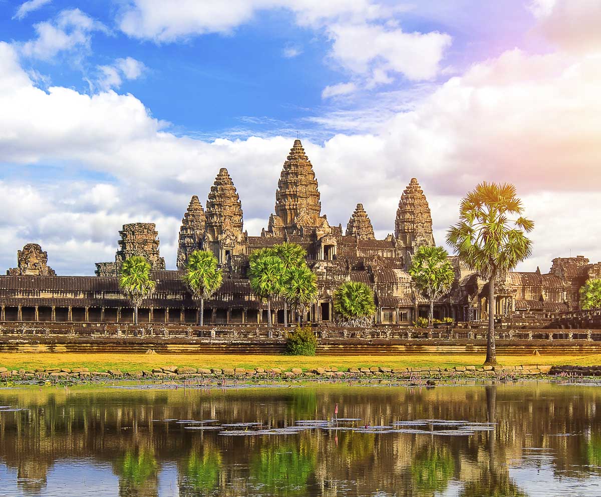 Templo Angkor Wat com seus cinco pináculos refletidos em lago calmo e céu azul.