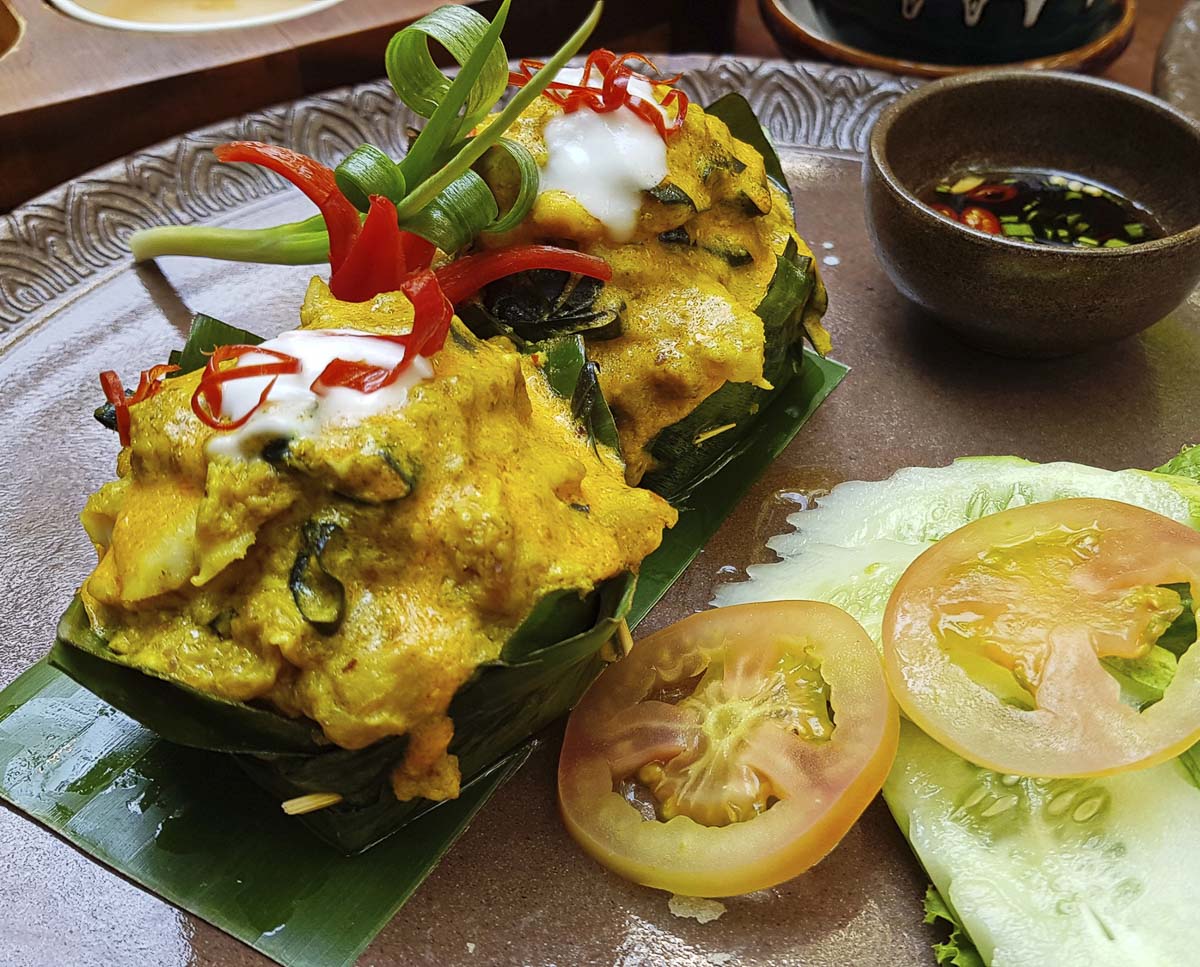Porção de fish amok, prato cambojano com molho amarelo cremoso servido em folha de banana, com tomate e pepino ao lado.