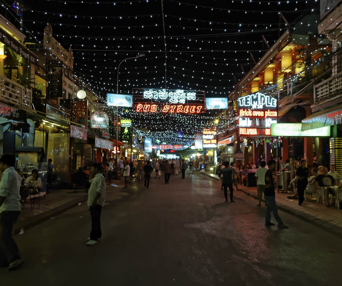 Rua iluminada com letreiros coloridos, bares e pessoas caminhando à noite em Siem Reap.
