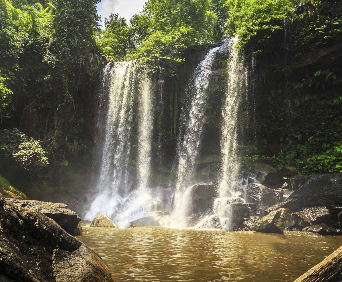 Cachoeira fluindo em três quedas entre rochas e vegetação densa, formando um poço de água.