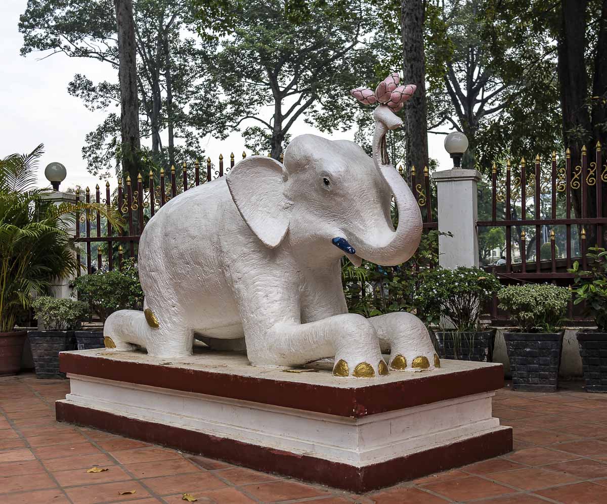 Escultura de elefante branco ajoelhado com flor de lótus na tromba em um jardim.