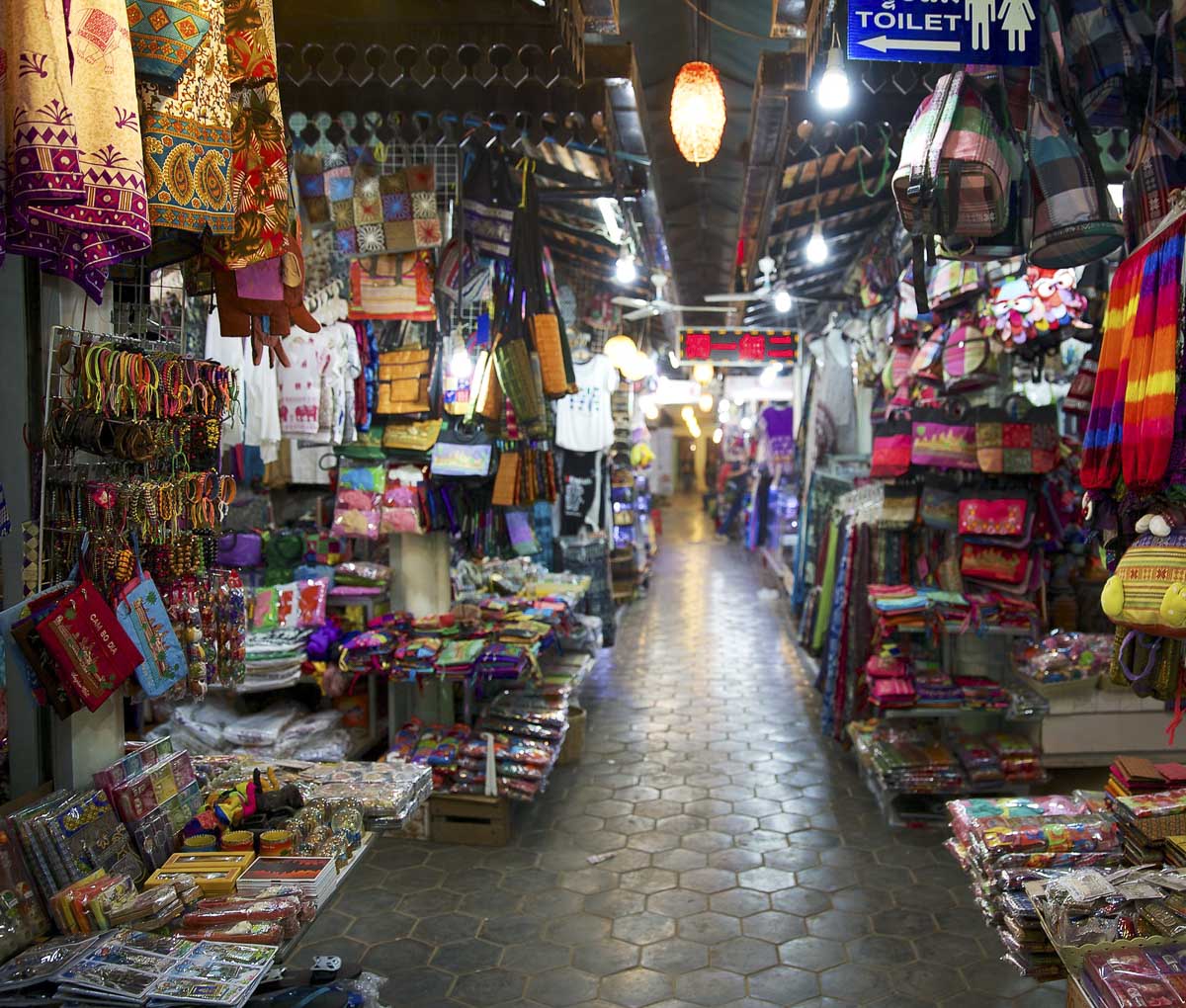 Corredor estreito com barracas coloridas vendendo bolsas, roupas e souvenires em Siem Reap.