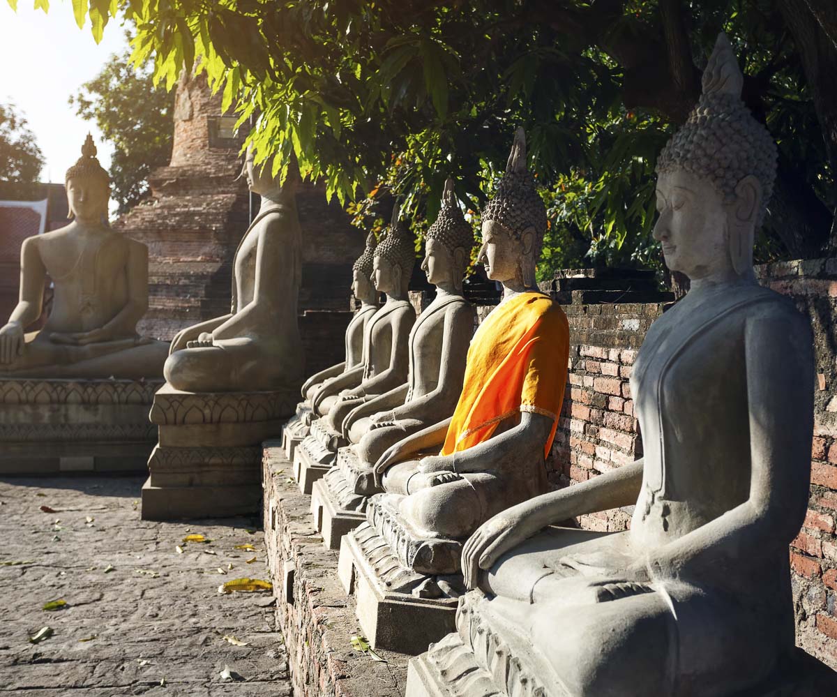 Estátuas de Buda sentadas em fila, com uma delas vestida com túnica laranja, em um templo ao ar livre na Tailândia.
