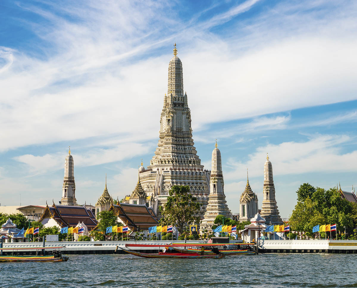 Vista frontal do templo Wat Arun, em Bangkok, com torres ornamentadas, bandeiras coloridas e barcos tradicionais no rio Chao Phraya.