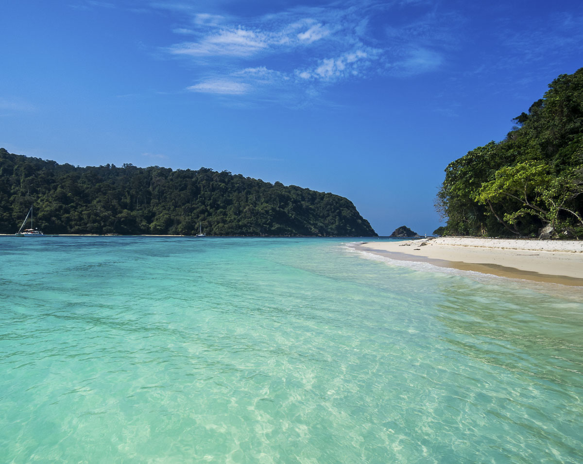 Praia deserta com águas cristalinas azul-turquesa, cercada por vegetação densa e colinas na Tailândia.