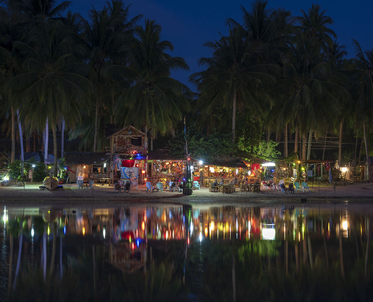 Bar de praia iluminado à noite com luzes coloridas refletidas na água, cercado por palmeiras altas na Tailândia.