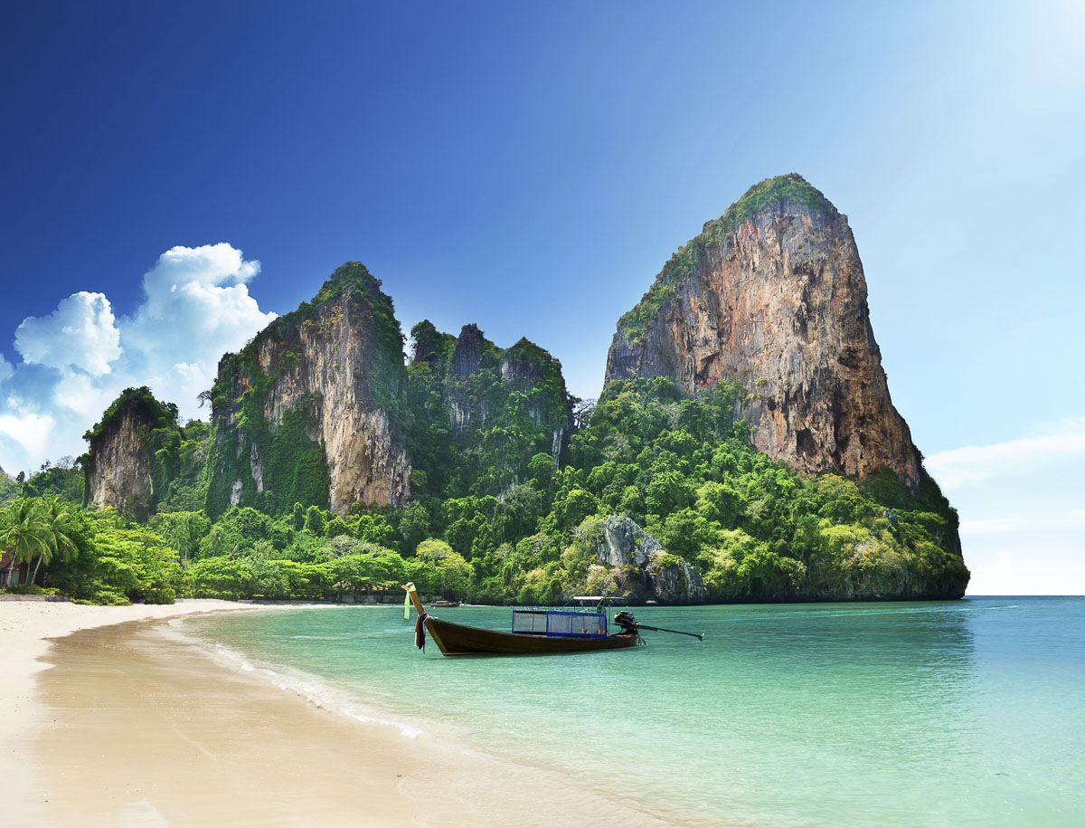Praia de areia clara com mar calmo, paredões rochosos cobertos de vegetação e um barco tradicional tailandês long-tail.