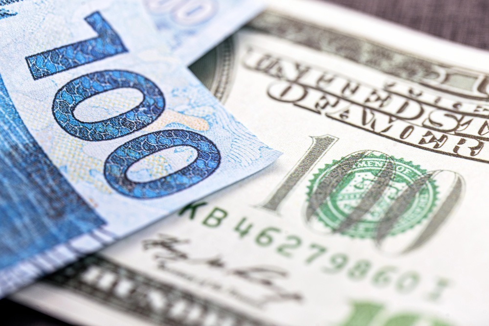 Notas de 100 reais e 100 dólares sobrepostas em close.