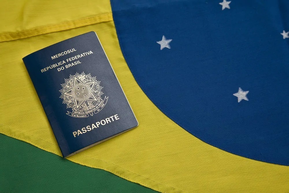 Passaporte azul do Brasil repousa sobre uma bandeira nacional nas cores verde, amarela e azul.