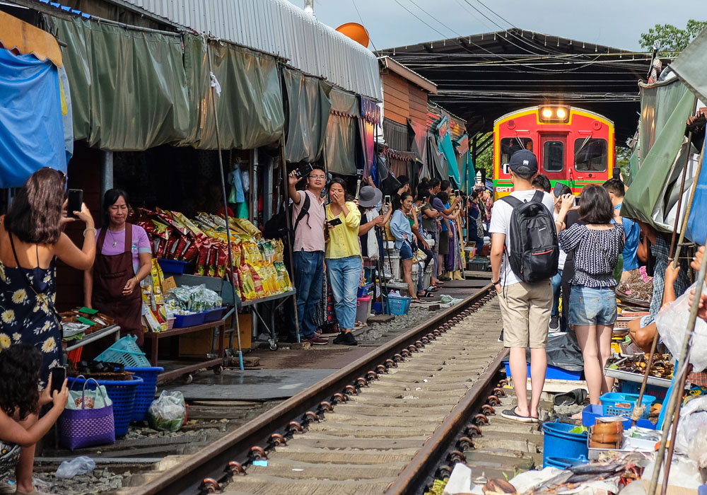 Trem avança devagar entre barracas e visitantes no Maeklong Railway Market, com céu colorido ao entardecer.