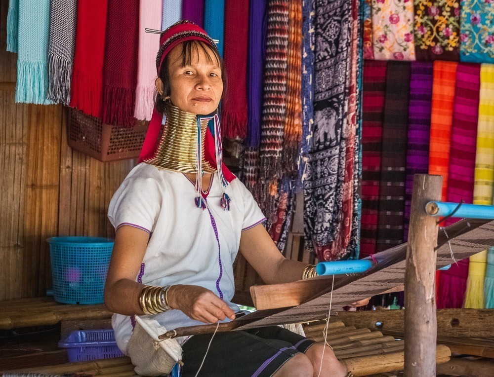 Mulher da tribo Karen tece em tear manual com tecidos coloridos ao fundo.