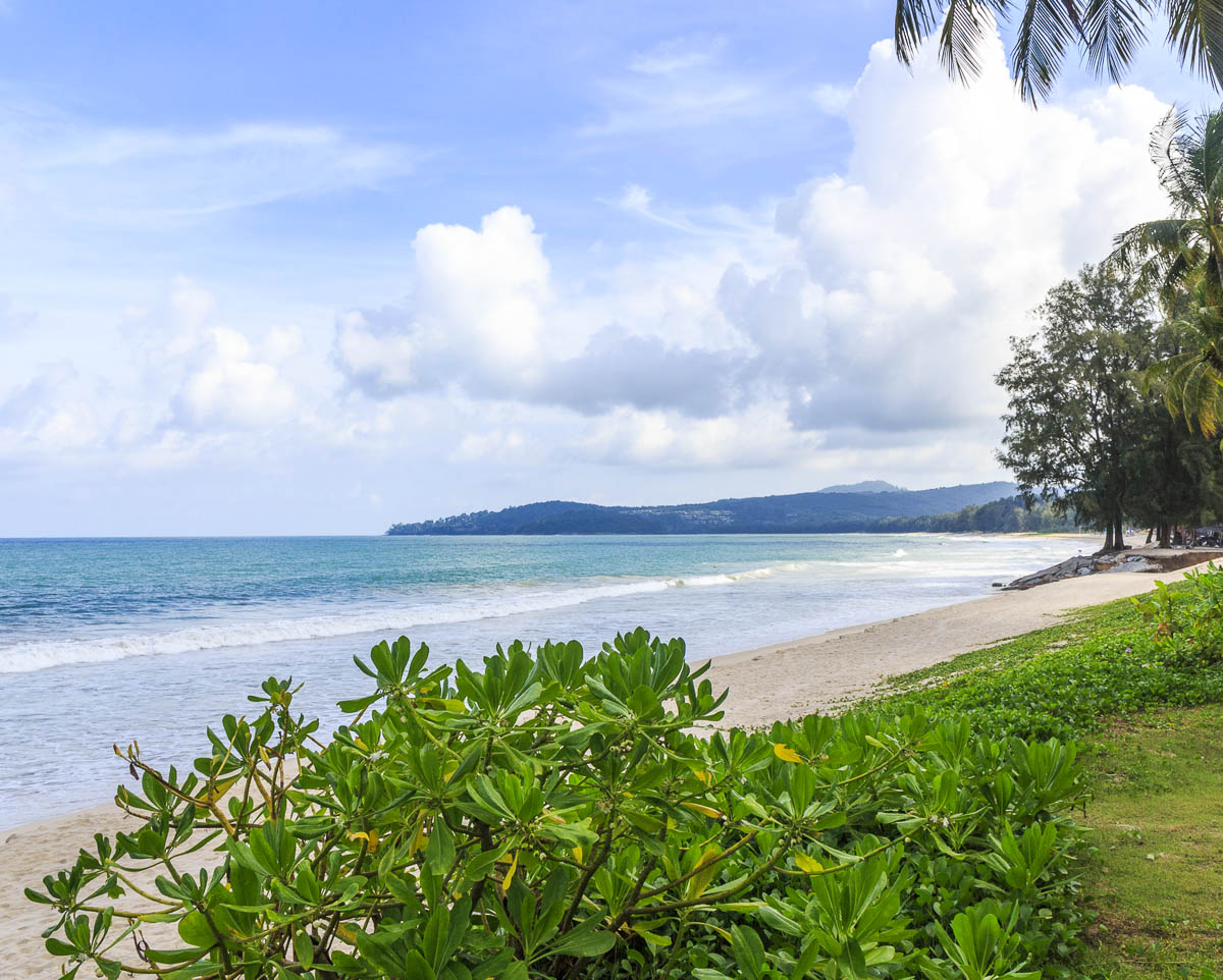 Praia tranquila na Tailândia com mar azul, vegetação tropical e céu parcialmente nublado.