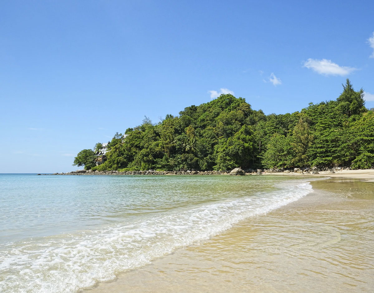 Praia deserta com mar calmo e águas cristalinas cercada por vegetação tropical na Tailândia.