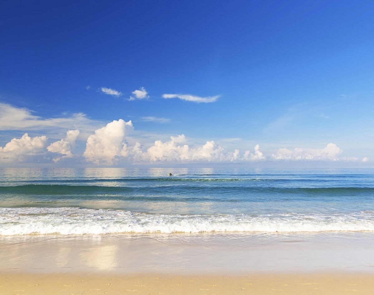 Praia tranquila com mar azul e céu com nuvens refletidas na água, em um cenário paradisíaco tailandês.