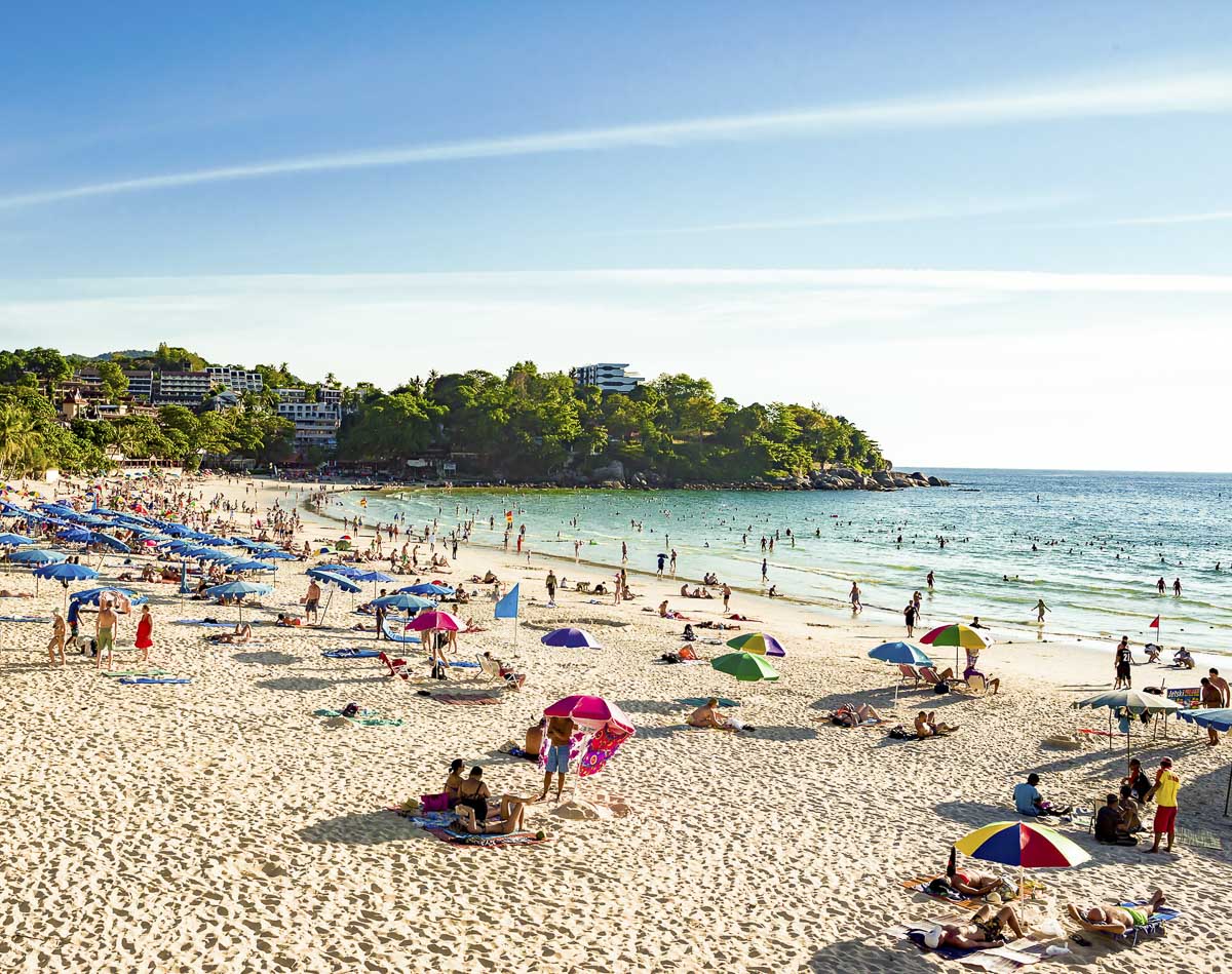 Praia lotada na Tailândia com guarda-sóis coloridos, turistas na areia e no mar, e construções ao fundo.
