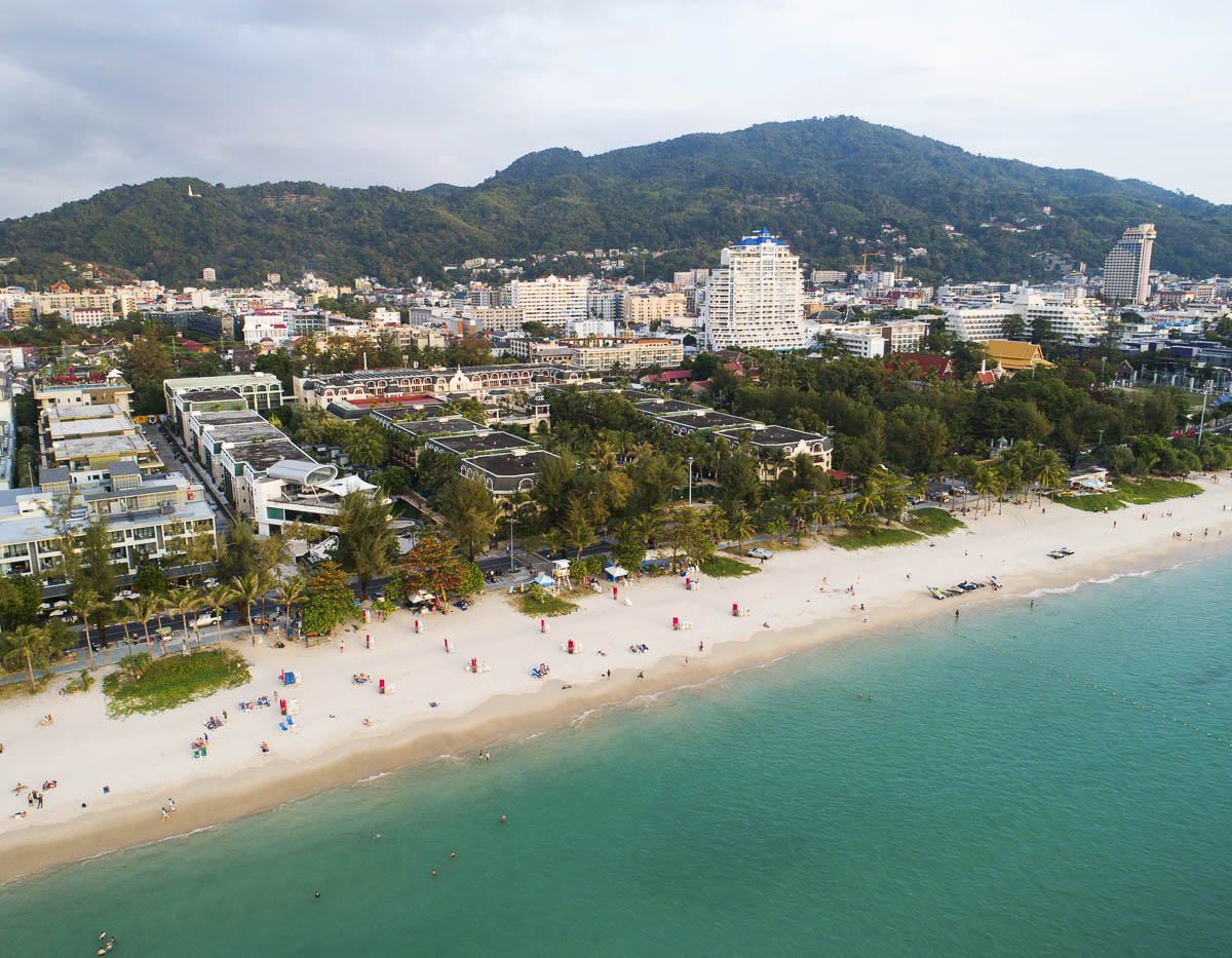 Vista aérea da praia de Patong na Tailândia com mar azul, faixa de areia clara e cidade entre colinas verdes.