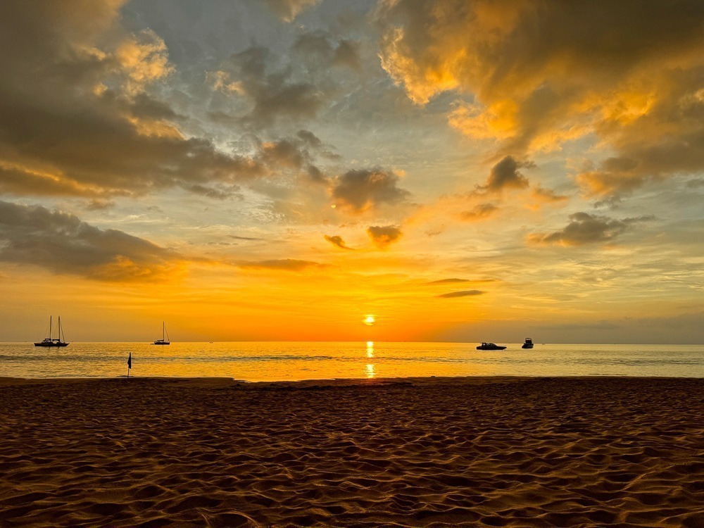 Fim de tarde dourado em Kantiang Bay, com o céu alaranjado refletido no mar calmo e quatro barcos flutuando ao fundo.