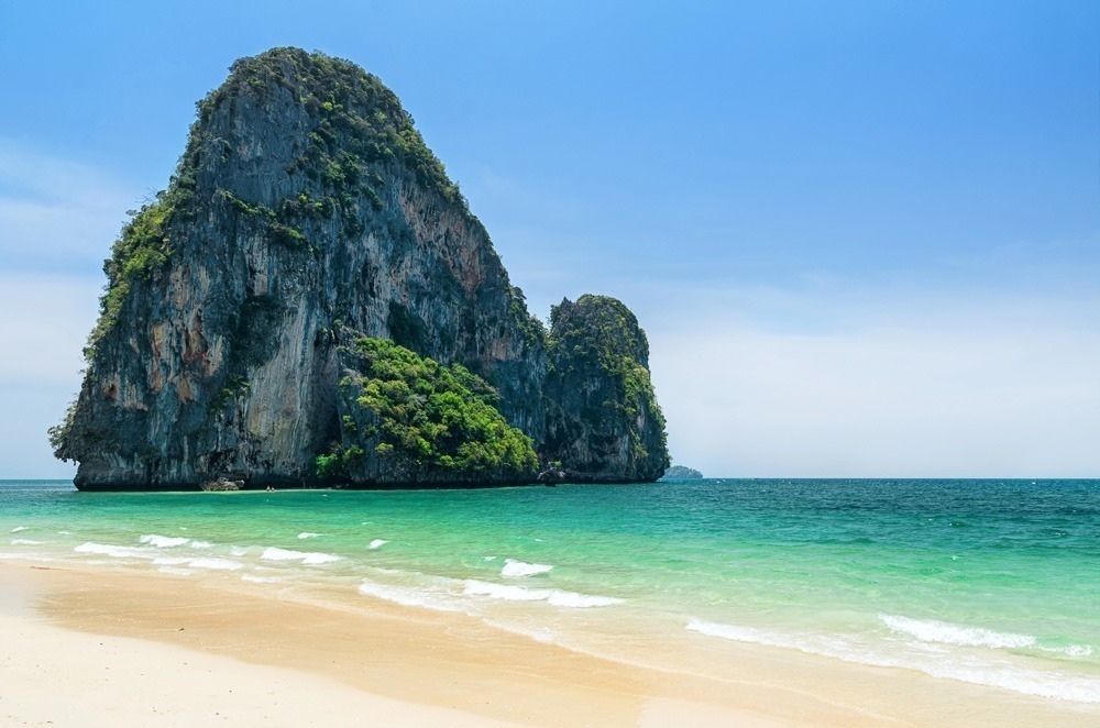 Praia de areia clara e mar esverdeado com o paredão rochoso de Phra Nang se erguendo próximo à orla.