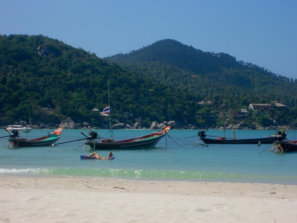 Areia clara, barcos longtail flutuando no mar e colinas cobertas de verde ao fundo em Thong Nai Pan.