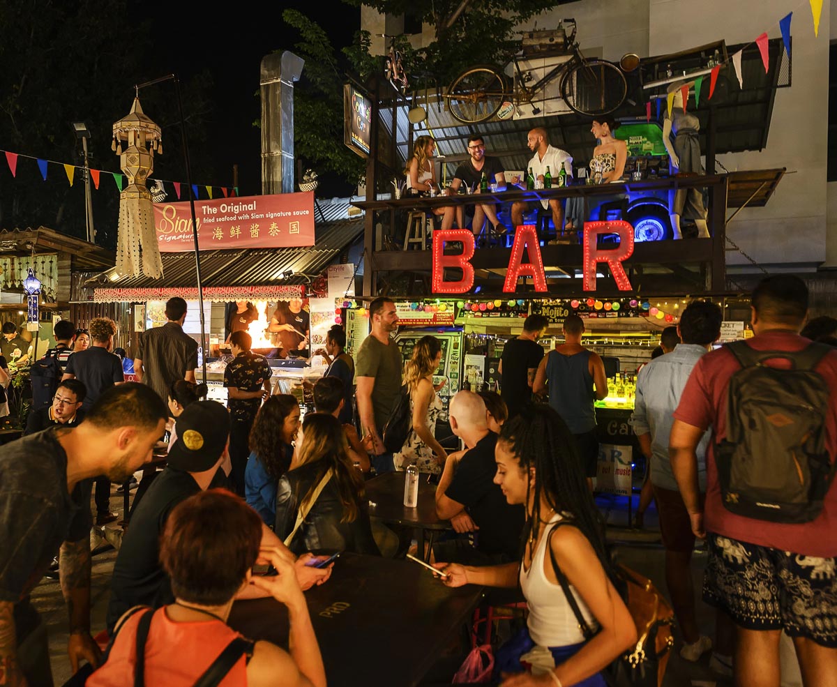Pessoas aproveitam a vida noturna de Chiang Mai em um mercado com bar iluminado e cheio de movimento.