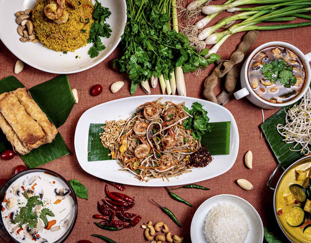 Pratos tailandeses variados dispostos em mesa com ingredientes frescos ao redor.