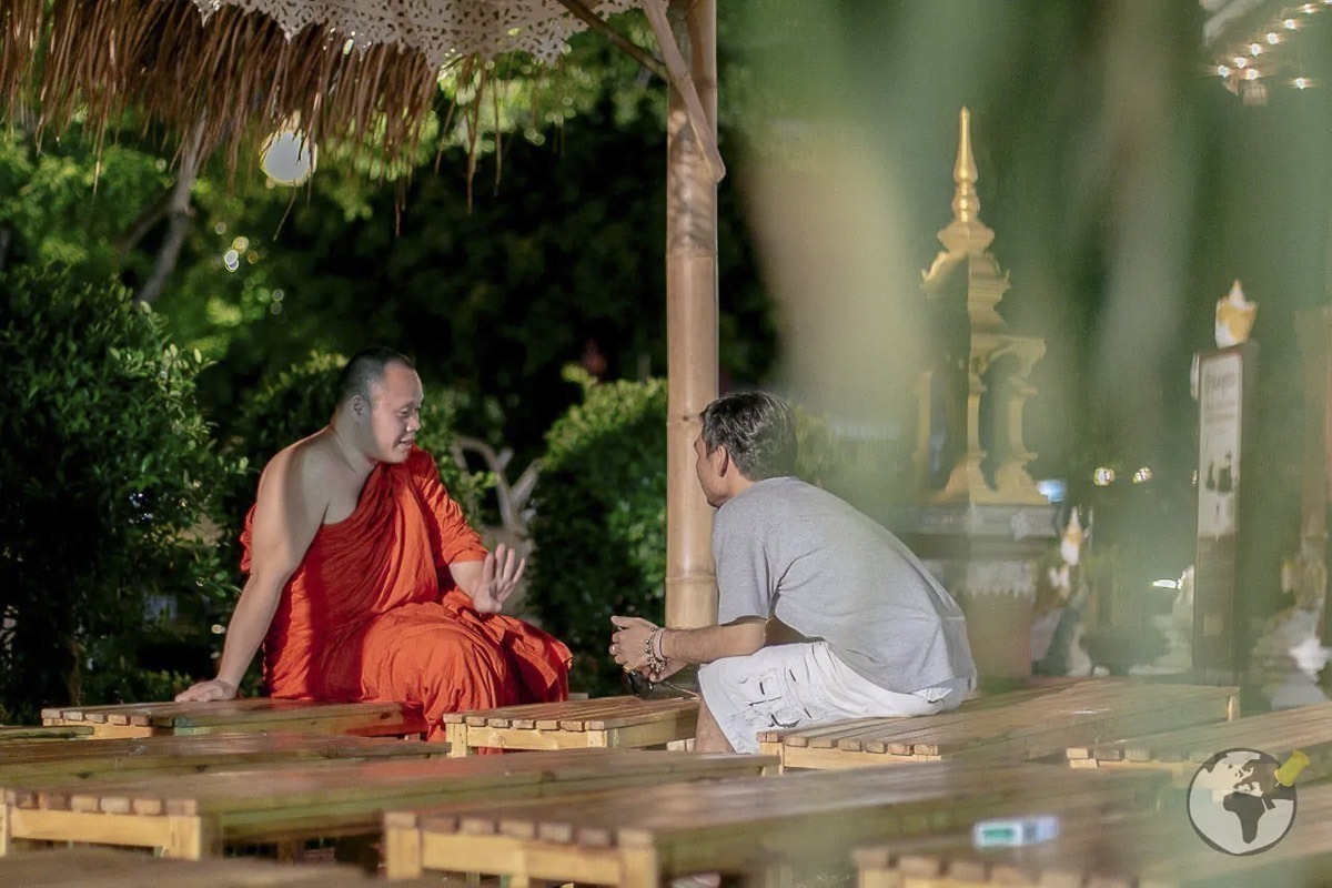 Monge budista conversa com visitante em área externa cercada por vegetação e lanternas.