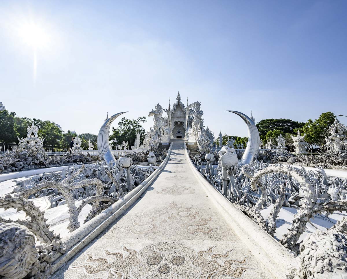Entrada do Wat Rong Khun com esculturas brancas detalhadas e passarela central que leva ao templo principal.