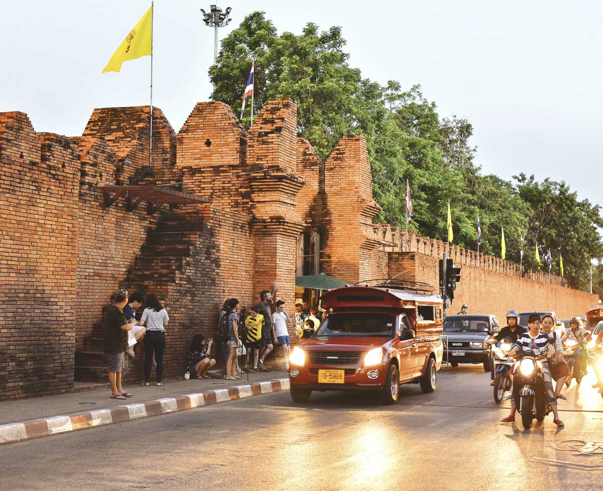 Trânsito movimentado e turistas em frente ao Portão Tha Phae, com muralha de tijolos antigos ao fundo.