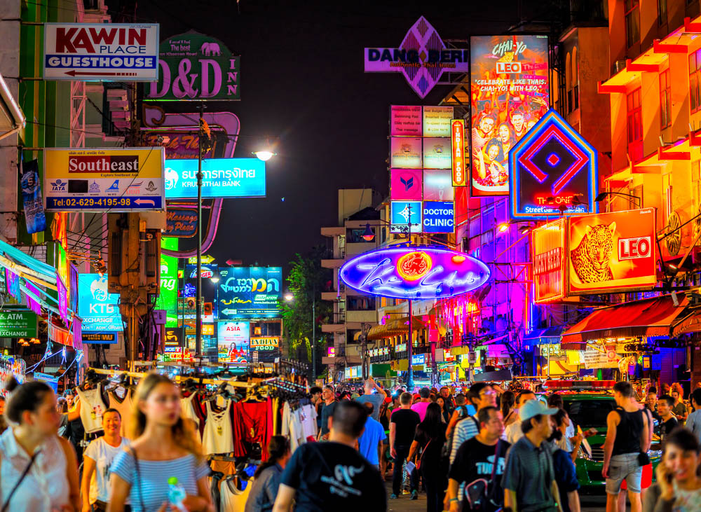 Rua Khao San movimentada à noite, iluminada por letreiros de néon e cheia de turistas.