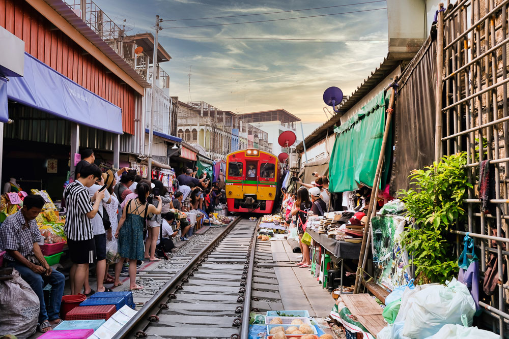 Trem vermelho e amarelo avança sobre trilhos rodeados por barracas e turistas no mercado de Maeklong.