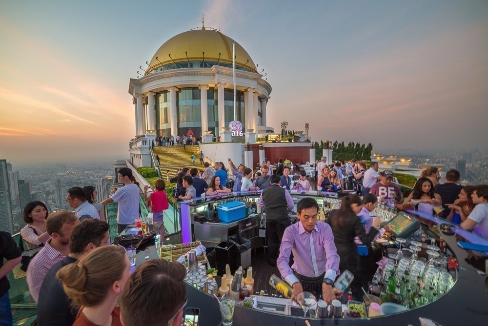 Sky Bar no topo do Lebua at State Tower, com pessoas bebendo ao entardecer e vista panorâmica de Bangkok.