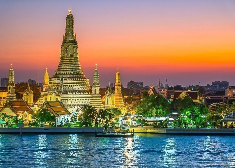 Wat Arun iluminado ao entardecer, com reflexo no rio Chao Phraya e céu colorido ao fundo.