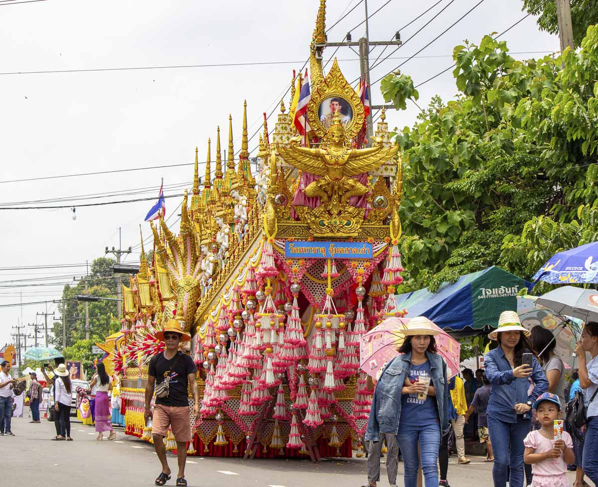 Desfile de rua com carros alegóricos dourados decorados e participantes vestindo roupas tradicionais durante o Festival Boon Bang Fai, na Tailândia.
