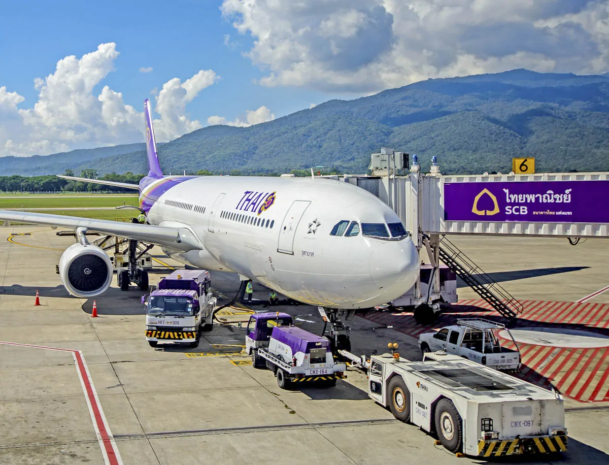 Avião da Thai Airways estacionado no aeroporto, com montanhas ao fundo, na Tailândia.