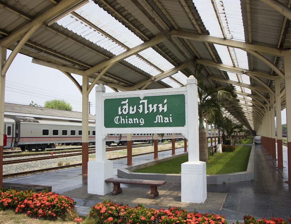 Placa da estação de trem de Chiang Mai com letreiro verde e trens ao fundo.