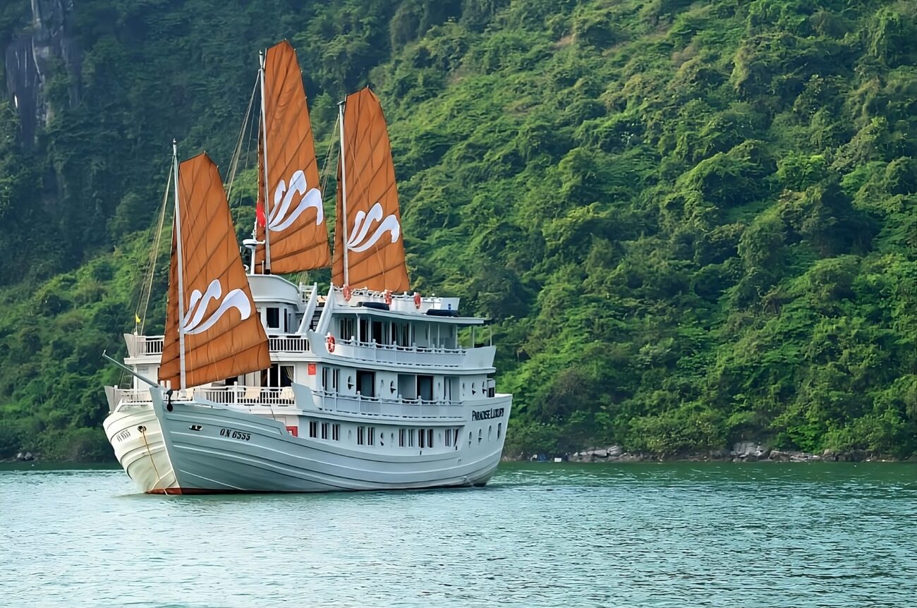 Barco de cruzeiro com velas laranja navega nas águas calmas de Halong Bay.
