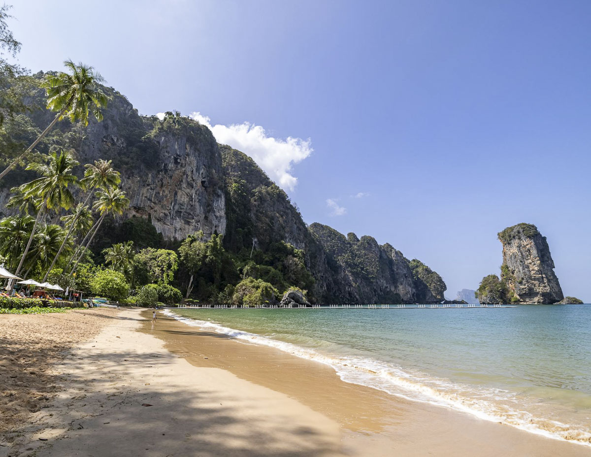 Praia de Ao Nang com mar calmo, coqueiros e paredões rochosos ao fundo, em Krabi.