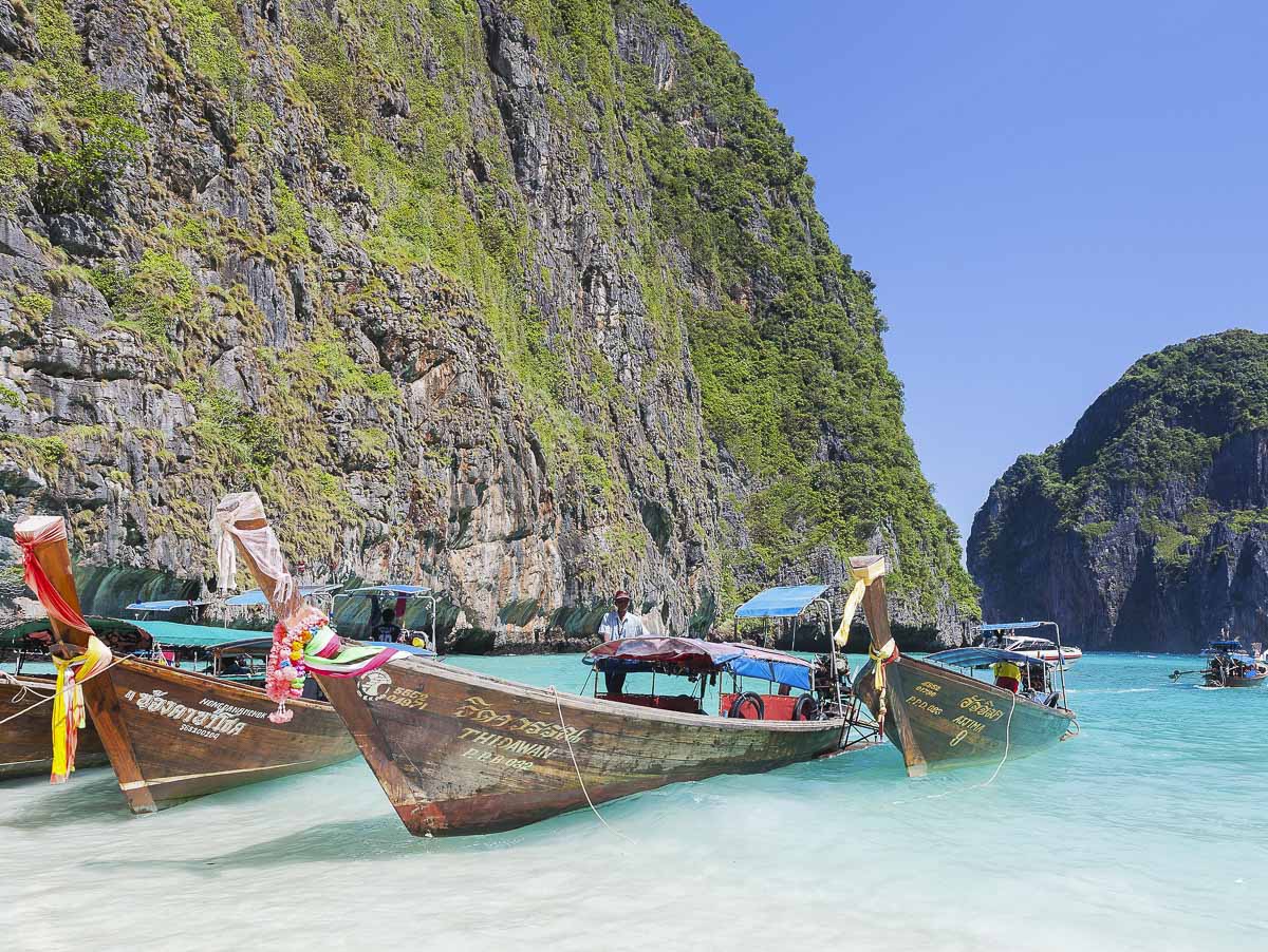 Barcos longtail coloridos ancorados nas águas turquesa de Maya Bay, cercada por falésias.