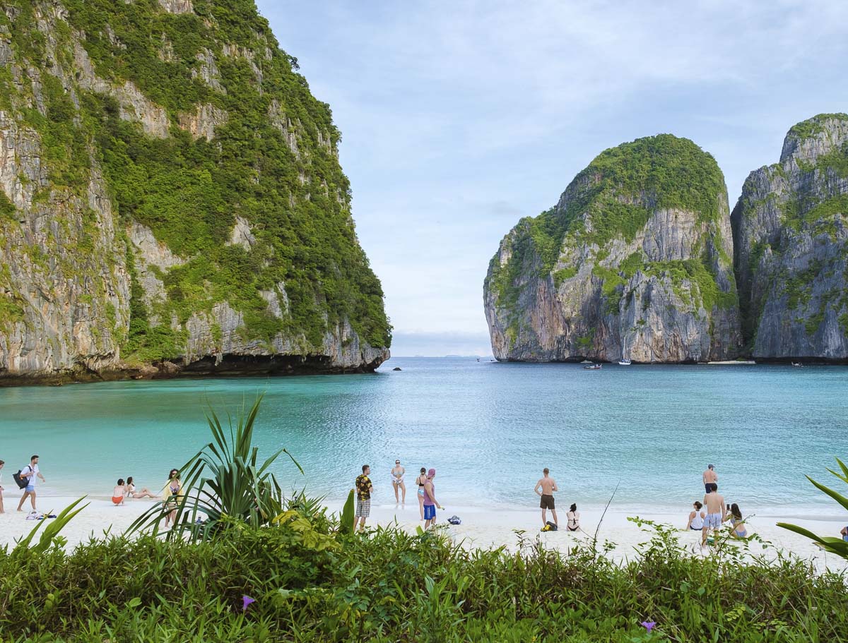 Turistas caminhando na areia branca de Maya Bay cercada por paredões verdes e mar azul.