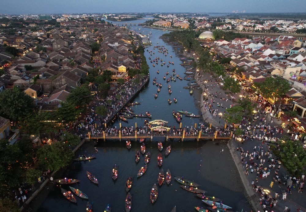 Vista aérea do rio Thu Bon em Hoi An, com barcos iluminados e pontes cheias de visitantes.