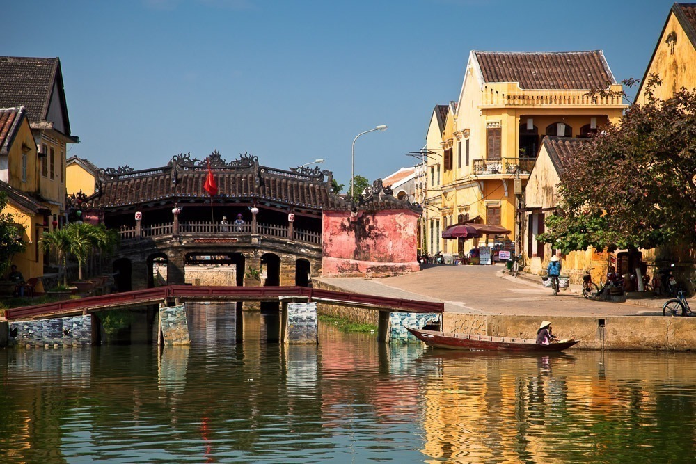 A Ponte Japonesa de Hoi An refletida nas águas do canal em um dia ensolarado.