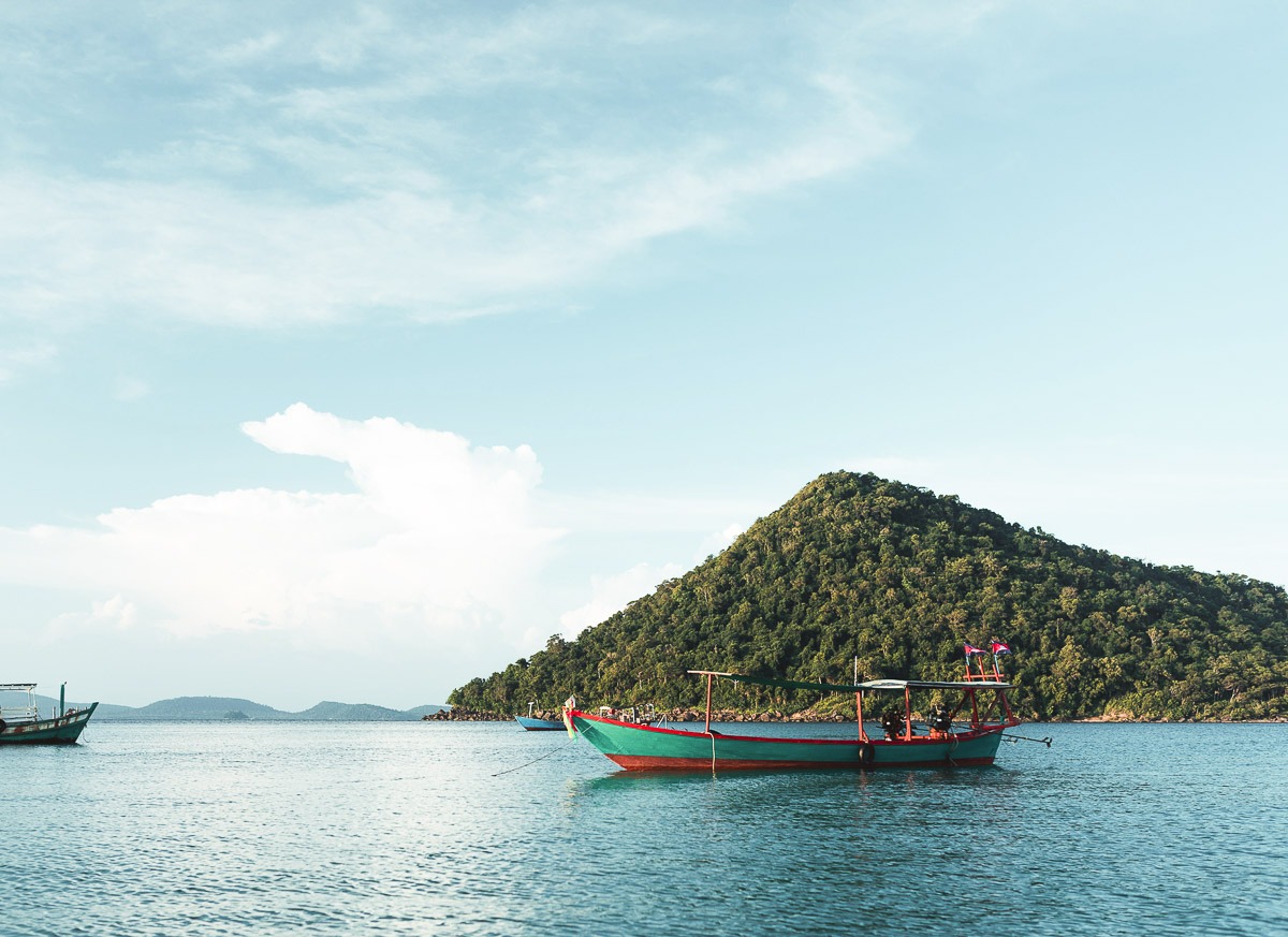 Barco de madeira colorido no mar calmo de Koh Rong, com colina verde ao fundo.