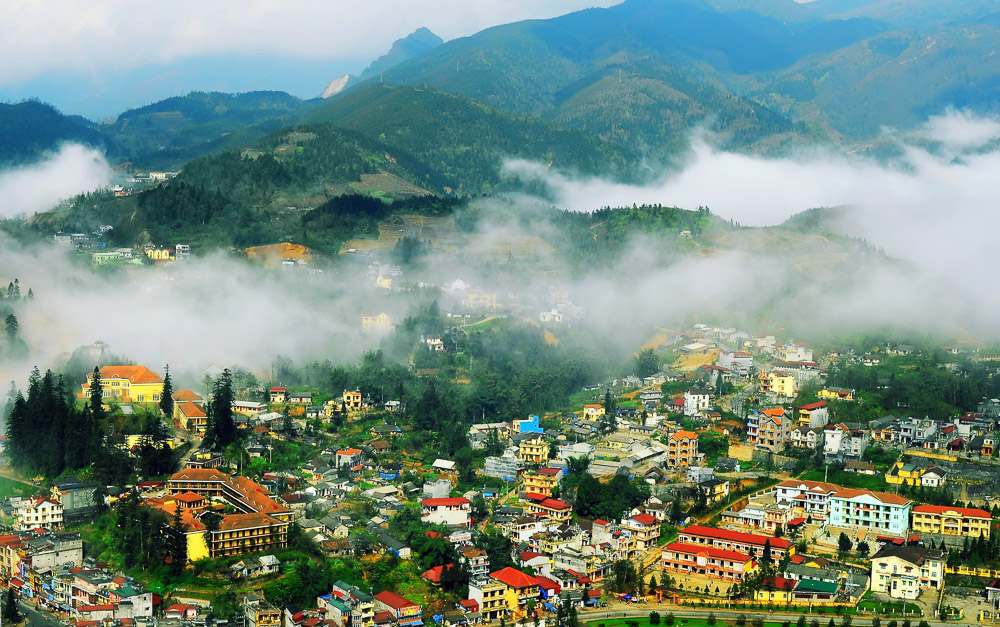Vista elevada de Sapa com névoa entre casas e montanhas ao fundo.