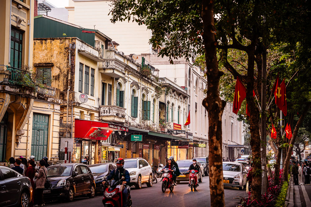 Rua movimentada do French Quarter em Hanói, com prédios coloniais e bandeiras vietnamitas.