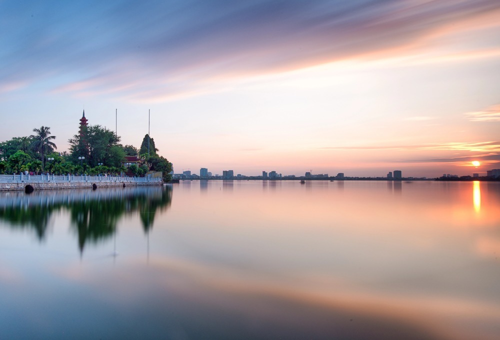 Pôr do sol sobre o West Lake em Hanói, com reflexos coloridos na água e pagode ao fundo.