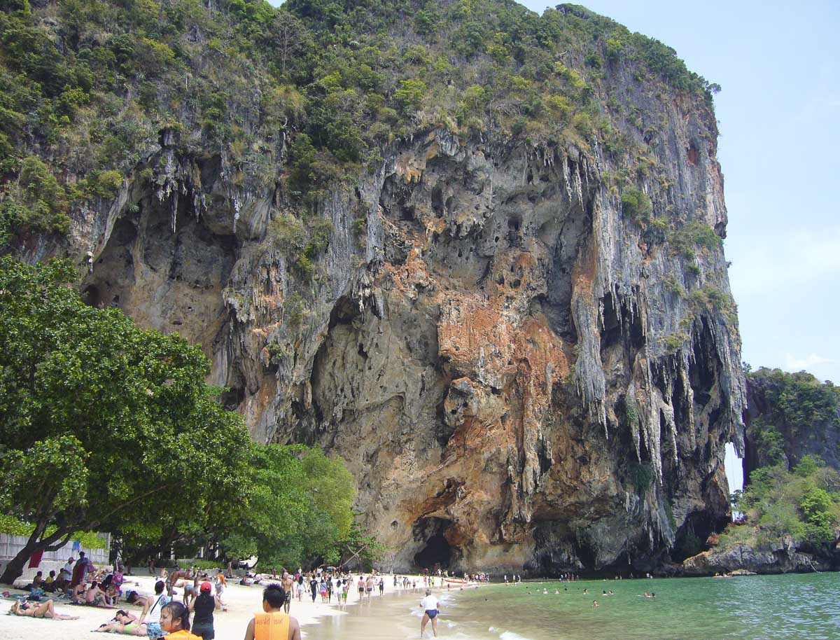 Entrada da caverna Phra Nang, com paredão rochoso e turistas na areia, em Railay, Tailândia.