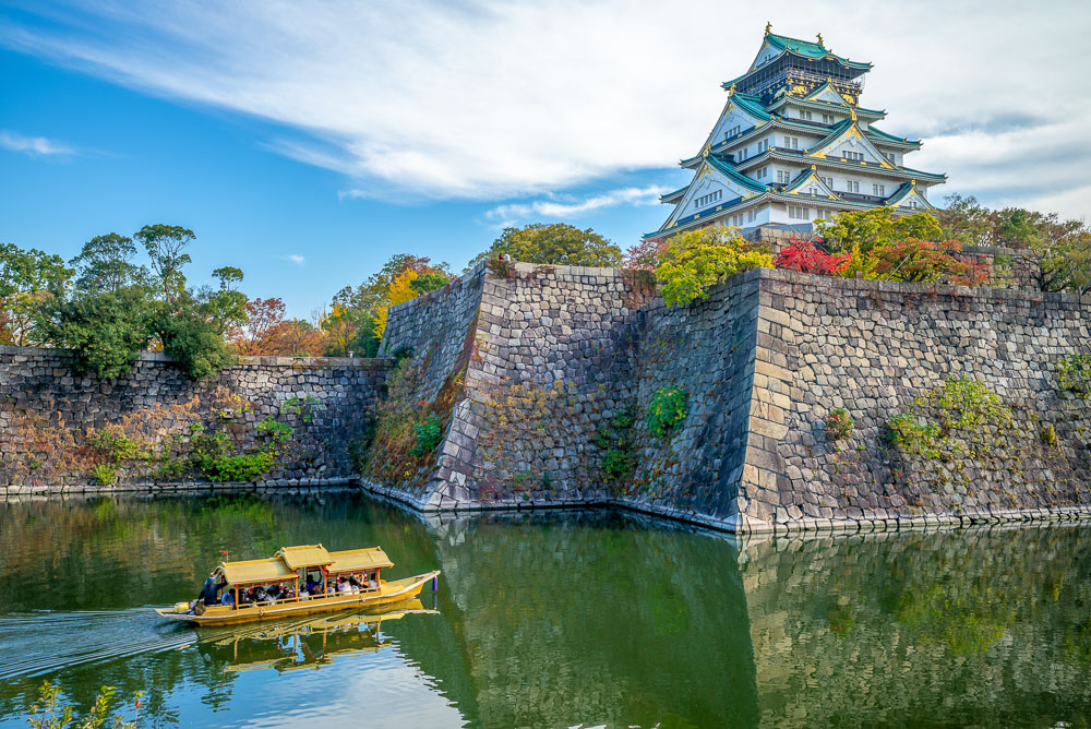 Barco cruzando o inner moat do Castelo de Osaka, com muralhas de pedra e o castelo no alto.