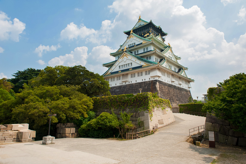 Torre principal do Castelo de Osaka vista a partir do caminho de acesso, com muralhas de pedra e vegetação ao redor.