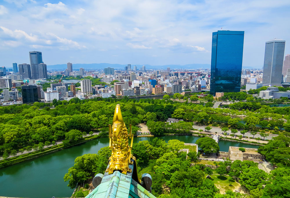Vista do alto do Castelo de Osaka com o fosso, áreas verdes e prédios modernos da cidade ao fundo.