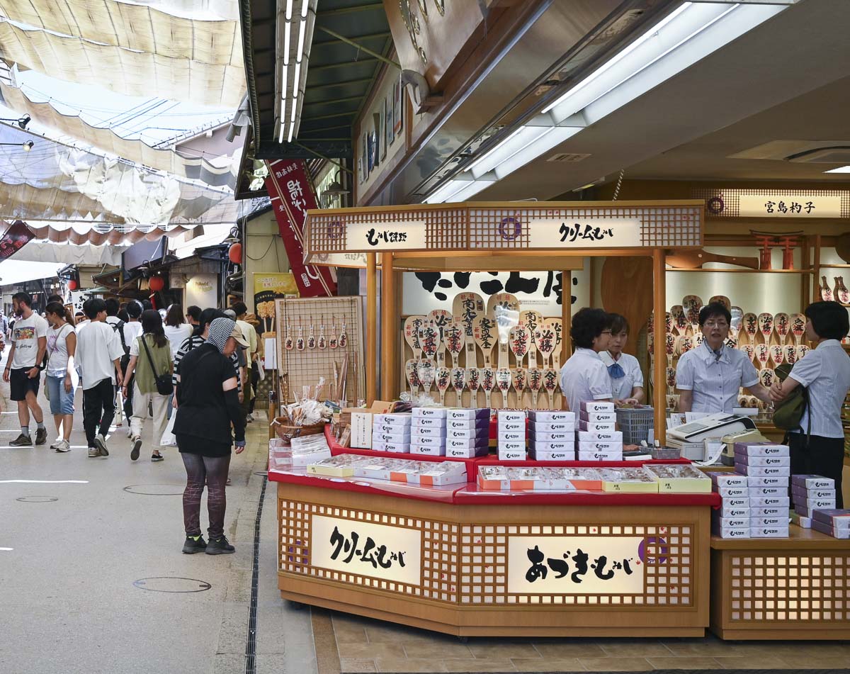 Rua comercial Omotesando, na ilha de Miyajima, com lojas tradicionais e visitantes caminhando.